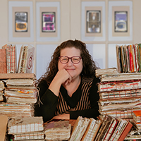Curly haired Laura Chenault sits with a smile surrounded by stacks of sketchbooks.