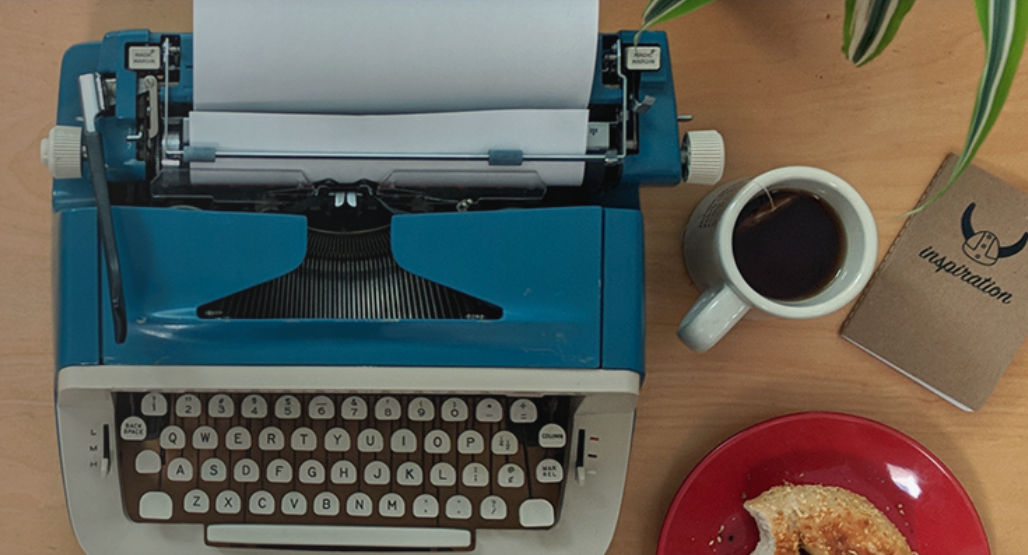 Picture of a blue and tan vintage typwriter with a cup of tea, a bitten bagel on a red plate, and a notebook that says inspiration.