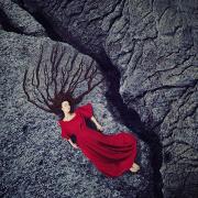 Self portrait by Kylli Sparre of a woman in a red dress laying on a rock next to a crack. Her hair is spread out and looks like tree roots.